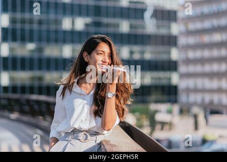 Horizontales Porträt einer attraktiven indischen Frau, die mit ihrem Smartphone eine Sprachnachricht sendet. Sie lehnt sich an das Geländer einer Brücke Stockfoto