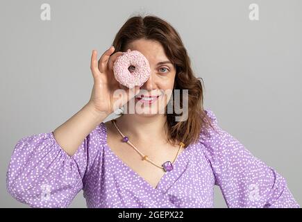 Mädchen hält einen Donut wie eine Brille. Das Mädchen lächelt in einem lila Kleid auf grauem Hintergrund. Stockfoto