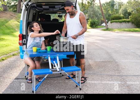 Ein junger tätowierter Mann, der in einer Campinggasküche über einem zusammenklappbaren Picknicktisch kocht, und seine Freundin schnappt sich Essen aus dem Topf. Stockfoto