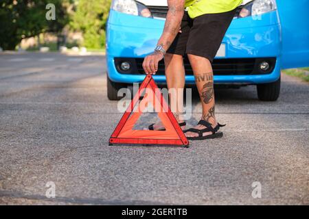 Nicht erkennbarer tätowierter Mann, der nach einer Panne in einer Notsituation ein reflektierendes Sicherheitsdreieck vor seinen Van legte. Stockfoto