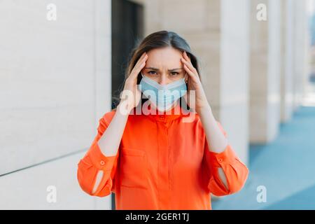 30-jährige Frau, die eine medizinische Maske trägt und an Migräne und Kopfschmerzen leidet Stockfoto