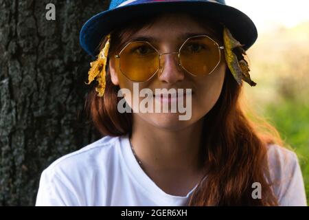 Unschärfe-Nahaufnahme-Porträt einer romantischen verträumten jungen Frau mit braunen Haaren, die einen Hut mit trockenen gelben Blättern und weißem T-Shirt im Freien trägt. Hallo Stockfoto