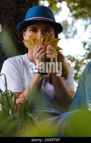 Unschärfe-Porträt einer lustigen jungen Frau mit braunen Haaren, die einen Hut im Freien trägt. Weibliche Hand mit gelben trockenen Blättern und bedecktem Gesicht. Hallo Herbst Stockfoto