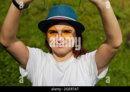 Unschärfe-Nahaufnahme Porträt einer glücklichen jungen Frau mit braunen Haaren trägt einen Hut im Freien. Gelbe Spiegelsonnenbrille auf dem Gesicht. Grüne Natur Hintergrund Stockfoto