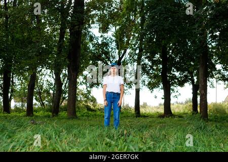 Unschärfe-Gesamtporträt einer ernsthaften jungen Frau mit braunen Haaren, die einen Hut im Freien trägt. Grüne Natur Bäume Hintergrund, Wald. Blick auf die Kamera Stockfoto