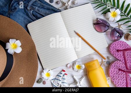 Konzept für Sommerferien. Strohhut, offenes Notizbuch und Strandzubehör mit Muscheln und Seesternen auf weißem Holzhintergrund Stockfoto