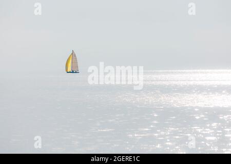 Minimalistisches Bild eines einzigen Segelbootes auf einem See an einem neblig bewölkten Tag mit nur einem Sonnenstrahl, der von der Wasseroberfläche reflektiert wird. Stockfoto