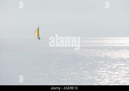 Minimalistisches Bild eines einzigen Segelbootes auf einem See an einem neblig bewölkten Tag mit nur einem Sonnenstrahl, der von der Wasseroberfläche reflektiert wird. Stockfoto