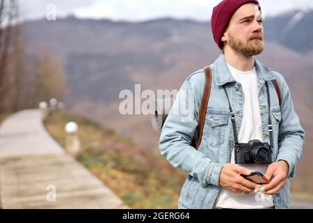 Reisender, der mit der Kamera in den Bergen unterwegs ist, die Landschaft genießt, Seitenansicht im Porträt. Bärtiger kaukasischer Rüde im lässigen Outfit im Freien, mit Rucksack. Stockfoto