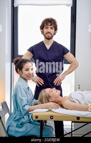 Zwei kaukasische Masseure üben das Massieren auf weiblichem Gesicht und Kopf des Klienten, männlicher und weiblicher Masseur in Uniform arbeiten zusammen, blonde Dame liegt auf massa Stockfoto