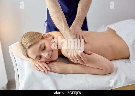 Junge blonde Frau erhält Rückenmassage von einem kurzhaarigen männlichen Masseur im Spa-Zentrum, entspannte kaukasische Dame genießt Spa-Behandlungen von professionellen, lyi Stockfoto