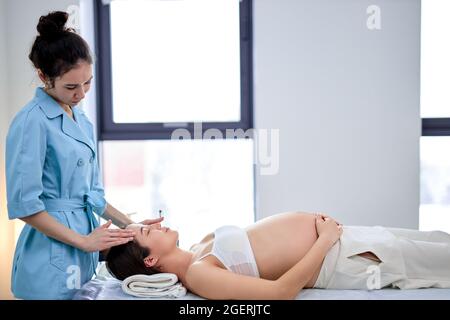 Seitenansicht Porträt eines jungen weiblichen Masseurs massiert Gesicht und Kopf des Schwangeren, lindert den Zustand während der Schwangerschaft, entspannt, in Spa-Kabine Stockfoto