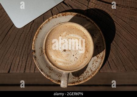 Tasse schwarzen Kaffee mit Milch in beige Handwerk Tasse auf einem massiven Holz Eiche Couchtisch mit Silber Laptop. Gemütlicher Raum für die Arbeit zu Hause während des COVID-19-Schlosses Stockfoto