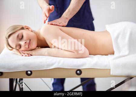 Wunderschöne Dame, die auf dem Bauch liegt, erhält eine Massage auf dem Rücken von einem männlichen Therapeuten, kaukasischer Typ in blauer Uniform, der die Rückenmuskeln von cl sorgfältig massiert Stockfoto