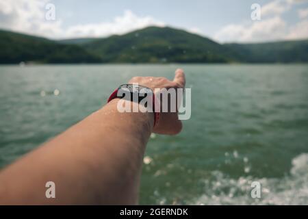 Selektive Fokussierung auf eine rote Laufuhr mit Wassertropfen auf dem Bildschirm und nasser Männerhand mit Finger, der auf die Berge über den See zeigt Stockfoto