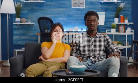 Junge interracial paar Fernsehen essen Popcorn im Wohnzimmer. Verheiratete, multiethnische Menschen, die die Kamera anschauten und einen Imbiss genossen und entspannende Aktivitäten genossen. Liebhaber gemischter Rennen Stockfoto