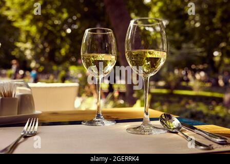 Zwei Gläser Weißwein, der in der Sonne funkelt, auf einem Restauranttisch mit einem Stadtgarten im Hintergrund. Stockfoto