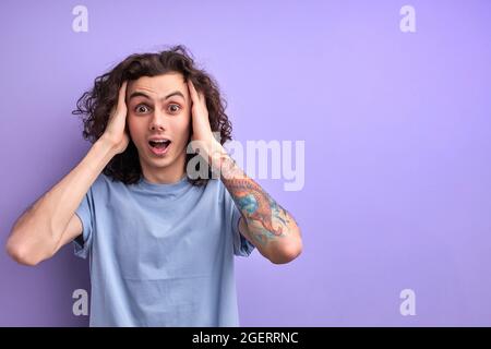 Mann mit lockigen Haaren ist schockiert, Überraschung und erstaunt Ausdruck mit Händen auf Gesicht isoliert über lila Studio Hintergrund, Porträt Stockfoto
