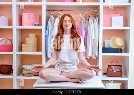 Positive glückliche junge Rothaarige Frau, die in einem aufgeräumten Raum voller Kleidung sitzt und meditiert, zu Hause, ruhig bleibt, in Yoga-Pose auf dem Tisch, die Hände namaste hält. N Stockfoto