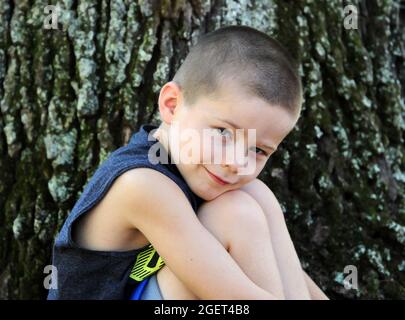 Kleiner Junge sitzt neben einem Baum im Freien. Er lehnt seinen Kopf gegen seine Knie, während er gerne draußen ist. Stockfoto