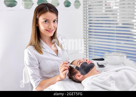 Confident Professionelle kaukasische Kosmetologin, die Bürstenmaske auf das hübsche schöne Gesicht des Kunden anwendet. Seitenansicht. Konzept der professionellen Kosmetologie A Stockfoto