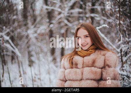 Attraktive junge Brünette Frau trägt Pelzmantel mit Schal, allein vor dem verschneiten Wald stehen, lächeln an der Kamera, während Sie Zeit im Freien verbringen o Stockfoto