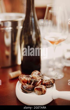 Vertikale Aufnahme von Escargots auf weißer Keramikpfanne serviert neben Weißwein, köstlichen gekochten Schnecken, genießen traditionelle französische Küche im Restaurant Stockfoto