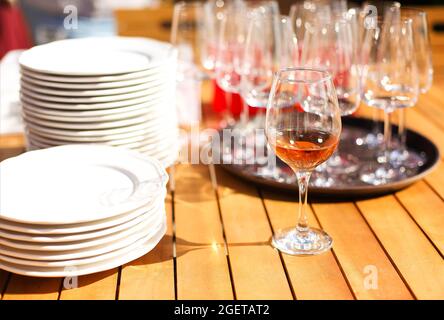 Holztisch mit vielen sauberen weißen Tellern und Weingläsern auf runden Tablett im Restaurant im Freien an sonnigen Tagen. Set aus Geschirr und Gläsern, auswählen Stockfoto