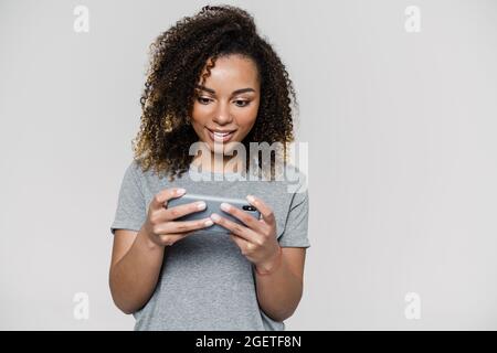 Eine glückliche Frau in einem grauen T-Shirt, die in das Telefon schaut und es in der Hand hält, während sie im Lichtstudio steht Stockfoto