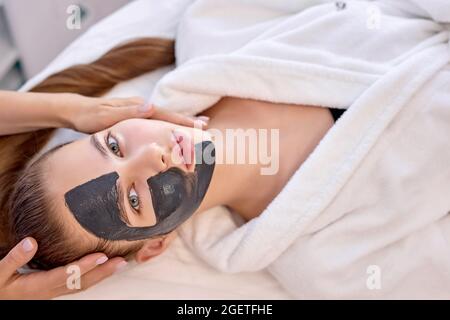 Junge kaukasische Frau nutzt die Dienste der professionellen Kosmetikerin bei at Spa. Cropped unkenntlich Meister Kosmetikerin setzt schwarze Maske auf Kundenfac Stockfoto