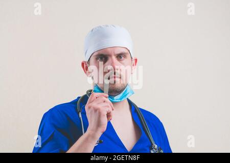 Junger Arzt Porträt in blauem Anzug, Stethoskop und schützende Atemmaske mit Spatel in der Hand in der Nähe seines Mundes aus der Nähe isoliert Stockfoto