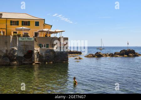 Eine Frau erfrischt sich an der Küste des kleinen Fischerdorfes, während Touristen auf der Terrasse eines Restaurants, Boccadasse, Genua, Italien, speisen Stockfoto