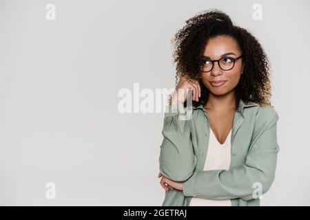 Eine nachdenkliche, lockige Frau in einer Brille, die zur Seite schaut und ihren Kopf mit der Hand aufstachelst, während sie im Studio steht Stockfoto