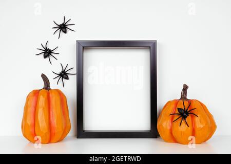 Schwarzer Rahmen mit Kürbis-Dekor auf einem Regal oder Schreibtisch. Halloween-Konzept. Porträtrahmen an einer weißen Wand mit Spinnen. Stockfoto