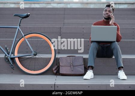 Beschäftigter junger Mann afrikanischer Herkunft mit Laptop, der im Freien auf dem Smartphone spricht Stockfoto