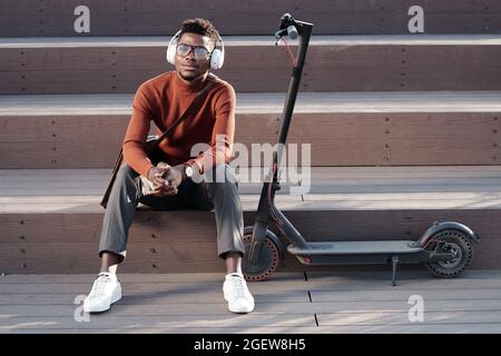 Fröhlicher schwarzer junger Mann, der Musik in Kopfhörern genießt, während er auf der Treppe sitzt Stockfoto