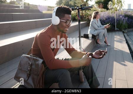 Glücklicher junger afrikanischer Mann mit Kopfhörern und Smartphone, der im Videochat im Freien kommuniziert Stockfoto