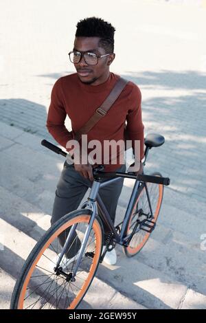 Junger Geschäftsmann mit Brillen und smarter Freizeitkleidung, der das Fahrrad schiebt, während er im Freien nach oben geht Stockfoto