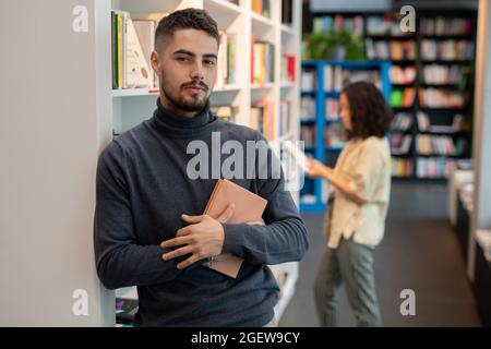 Bärtiger junger Mann mit Buch, das vor der Kamera steht, gegen weibliche Literatur, die sich für sie entscheidet Stockfoto
