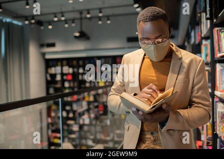 Junger Mann afrikanischer Herkunft liest Buch, während er in einem großen Buchladen steht Stockfoto