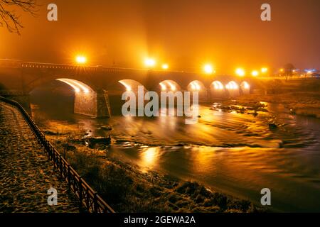 Herbstnachtsansicht einer alten Backsteinbrücke in Bögen gebaut, wo Nebel steigt aus dem Fluss alte Backsteinbrücke über einen breiten Fluss in einer dunklen Herbstnacht mit Stockfoto