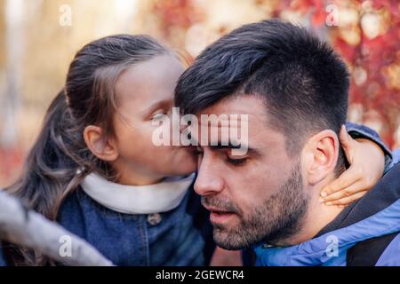 Nahaufnahme Mann und Mädchen Familienportrait. Ehrfürchtige dunkelhaarige Mann und flüsterte kleines Mädchen in blauer Jacke im Herbst Park Hintergrund Stockfoto