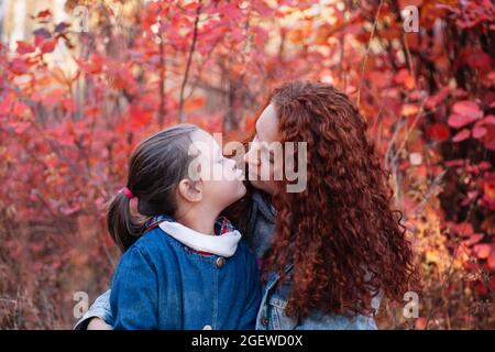 Nahaufnahme Liebeskonzept. Nettes kleines Mädchen küssen ihre Mutter mit langen kurvigen Haaren auf Herbst Wald Hintergrund Stockfoto