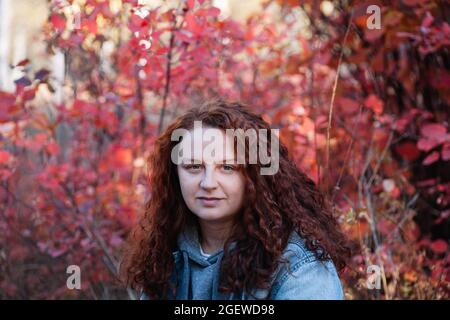 Frau Porträt aus nächster Nähe. Frau mit langen kurvigen braunen Haaren im Herbstwald mit rotem Busch auf dem Hintergrund Stockfoto