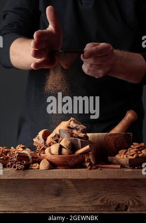 Der Koch streut die Schokolade mit Kakaopulver. Auf einem alten Holztisch Süßigkeiten, Kaffeebohnen, Zimt, Anis, Nüsse und Stücke von gebrochenem schwarzem Choco Stockfoto