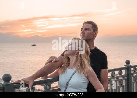 Ein Mann und eine Frau stehen umarmt auf dem Damm und schauen beim Sonnenuntergang auf das Meer. Generation der Millennials. Stockfoto