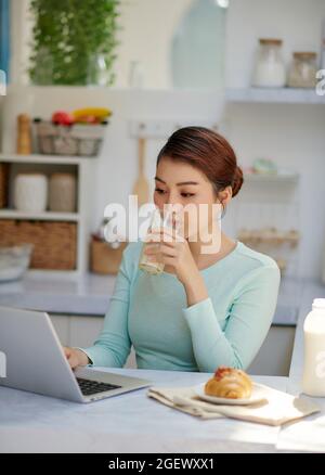 Attraktive Frau entspannend auf Ihrem Laptop und posieren beim Trinken ein Glas Milch in Ihrer Küche Stockfoto