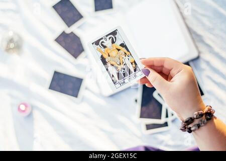 Weibliche Hand mit lila Nägeln hält Tarot-Karte namens der Teufel über der weißen Oberfläche mit offenem Notizbuch und Kerze. Draufsicht. Minsk, Weißrussland - 07.28.2021 Stockfoto