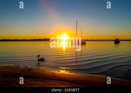 Die Boote ankerten in der Bribie Island Australia Bay bei Sonnenuntergang, während die Sonne über dem Horizont verschwindet, während Licht über dunkles Wasser auf das nahe Ufer und p ausläuft Stockfoto