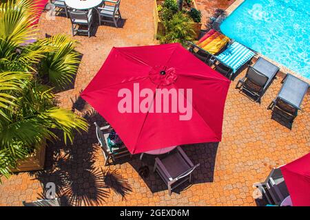 Strukturierte rote Sonnenschirme am Pool über Tischen und Stühlen neben Liegestühlen mit Strandhandtüchern und Topfpalmen auf der orangefarbenen Fliesenterrasse an sonnigen Tagen - top Stockfoto